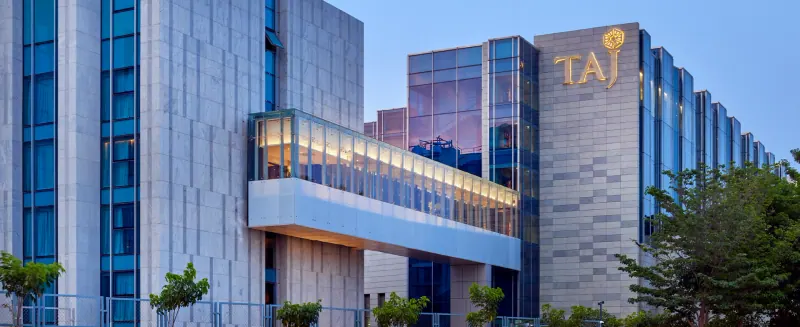 Dusk view of the Taj Bangalore expansion, with stone-clad towers, a glazed bridge lit from within and the Taj sign on the facade.