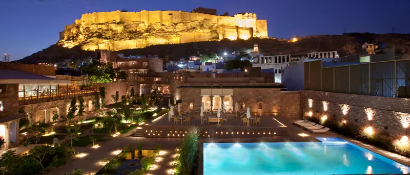 RAAS Jodhpur at night, with Mehrangarh Fort floodlit on the hill above and a lit courtyard and pool in the foreground.